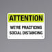 A black and yellow aluminum sign that says "Attention / We're Practicing Social Distancing"