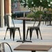 A Lancaster Table & Seating Excalibur square dining table with a textured wood finish and a cross base plate on a patio with black chairs.