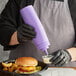 A person in gloves pouring sauce from a purple Choice wide mouth squeeze bottle onto a container of food.