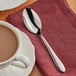 An Acopa Remy stainless steel teaspoon on a napkin next to a cup of coffee.