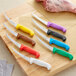 Choice 6" Curved Stiff Boning Knife with Yellow Handle next to a group of knives with different colored handles on a cutting board.