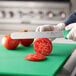 A person using a Choice serrated bread knife with a green handle to cut a tomato on a white rectangular object.
