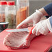 A person using a Choice narrow stiff boning knife with a red handle to cut meat on a red cutting board.