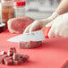 A person wearing gloves uses a Choice 6" Chef Knife to cut meat on a red cutting board.
