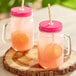 Two Acopa glass jars with pink liquid and pink metal lids with straws on a wood surface.