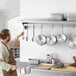 A man in a professional kitchen hanging pots and pans on a Regency stainless steel wall mounted pot rack.