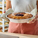 A person holding a round, deep pie with a golden crust and a fruit filling in the center, using oven mitts and a metal pie pan.