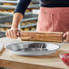 A person is rolling dough with a wooden rolling pin next to an empty round metal pie pan on a wooden countertop.