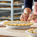 A person is crimping the edge of an unbaked pie with a decorative top crust in a round metal pie pan on a wooden surface.