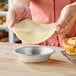 A person is holding a round piece of dough above a small, empty metal pie pan on a wooden surface.