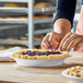 A person is shaping the crust of an unbaked fruit pie in a round metal pie pan on a wooden surface.