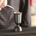 A person using an Arcoroc Ellis black steel bell jigger to pour liquid into a cup on a metal tray.