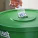 A hand putting a plastic bottle into a green Lavex recycling can with a round lid with a hole in it.