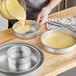 A person pouring yellow cake batter into a round metal cake pan.