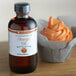 A bottle of LorAnn Oils All-Natural Orange Super Strength Flavor on a wooden table next to a cupcake with orange frosting.