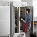 A man in a red apron and gloves opening the solid doors of a Beverage-Air Vista series reach-in freezer in a professional kitchen.