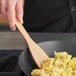 A person stirring scrambled eggs in a frying pan with an American Metalcraft wooden spatula.