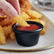 A person dipping a french fry into a black Acopa ramekin of ketchup.