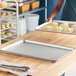 A perforated metal baking sheet is placed on a wooden countertop in a kitchen setting, with a person holding a tray of unbaked croissants in the background.