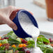 A hand pouring white dressing into a bowl of salad using a blue Acopa smooth melamine ramekin.