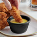 A person dipping a fried chicken strip into a black fluted ramekin of sauce.