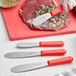 A Choice stainless steel sandwich spreader with a red handle being used to spread food on a cutting board.