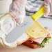 A person using a Choice stainless steel sandwich spreader with a neon yellow handle to spread a sandwich.