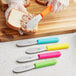 A gloved hand uses a Choice stainless steel sandwich spreader with a neon green handle to spread food on a sandwich.