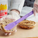 A person's hand with a Choice purple sandwich spreader spreading peanut butter on a bagel.