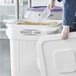 A person wearing gloves is scooping ingredients from a large white storage bin labeled 'BAKER'S LANE,' with the lid being held open.
