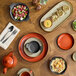 An orange porcelain coupe plate displayed on a wooden table set with various dishes and tableware.