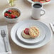 A white Acopa stoneware plate with a biscuit, jam, fruit, and a cup of coffee on it.