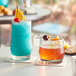 An Acopa rocks glass filled with liquid next to a blue drink with a cherry on top.