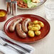 A plate of sausages and potatoes on an Acopa Sedona Orange stoneware coupe plate with a knife and fork.
