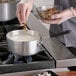 A person in a white coat cooking food in a Choice aluminum sauce pan on a stove.