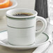 A Tuxton eggshell white china cup with a green stripe on a table with a cup of coffee and a spoon.