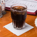 A glass of brown liquid with ice on a dove gray Hoffmaster beverage napkin.