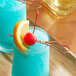 A blue drink with a cherry and a red bamboo heart skewer with a slice of orange.