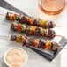 Grilled meat and vegetable skewers on a table with a cup of sauce.