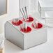 A stainless steel flatware organizer with red perforated plastic cylinders holding silverware.