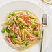 A plate of Barilla penne pasta with peas, ham, and vegetables with a fork and a glass of white wine.