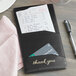 A black H. Risch padded check presenter with a credit card and thank you card and pen.