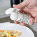 A hand holding a Tablecraft glass shaker with a white cap opening a bottle of salt.