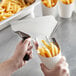 A hand using a Choice stainless steel French fry scoop to scoop French fries into a white container.