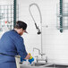 A man in a blue uniform using a Regency deck mount pre-rinse faucet to wash dishes in a professional kitchen sink.