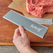 A hand holding a Schraf knife with a gray polypropylene blade guard over a piece of meat on a counter.