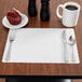 A customizable off-white paper placemat with a scalloped edge on a table with a white mug and muffin.
