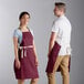 A man and woman wearing burgundy bib aprons with natural webbing accents.