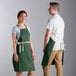 A man and woman wearing Choice hunter green poly-cotton aprons with natural webbing accents.