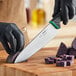 A person wearing black gloves uses a Schraf chef knife with a green handle to cut a purple potato on a counter.